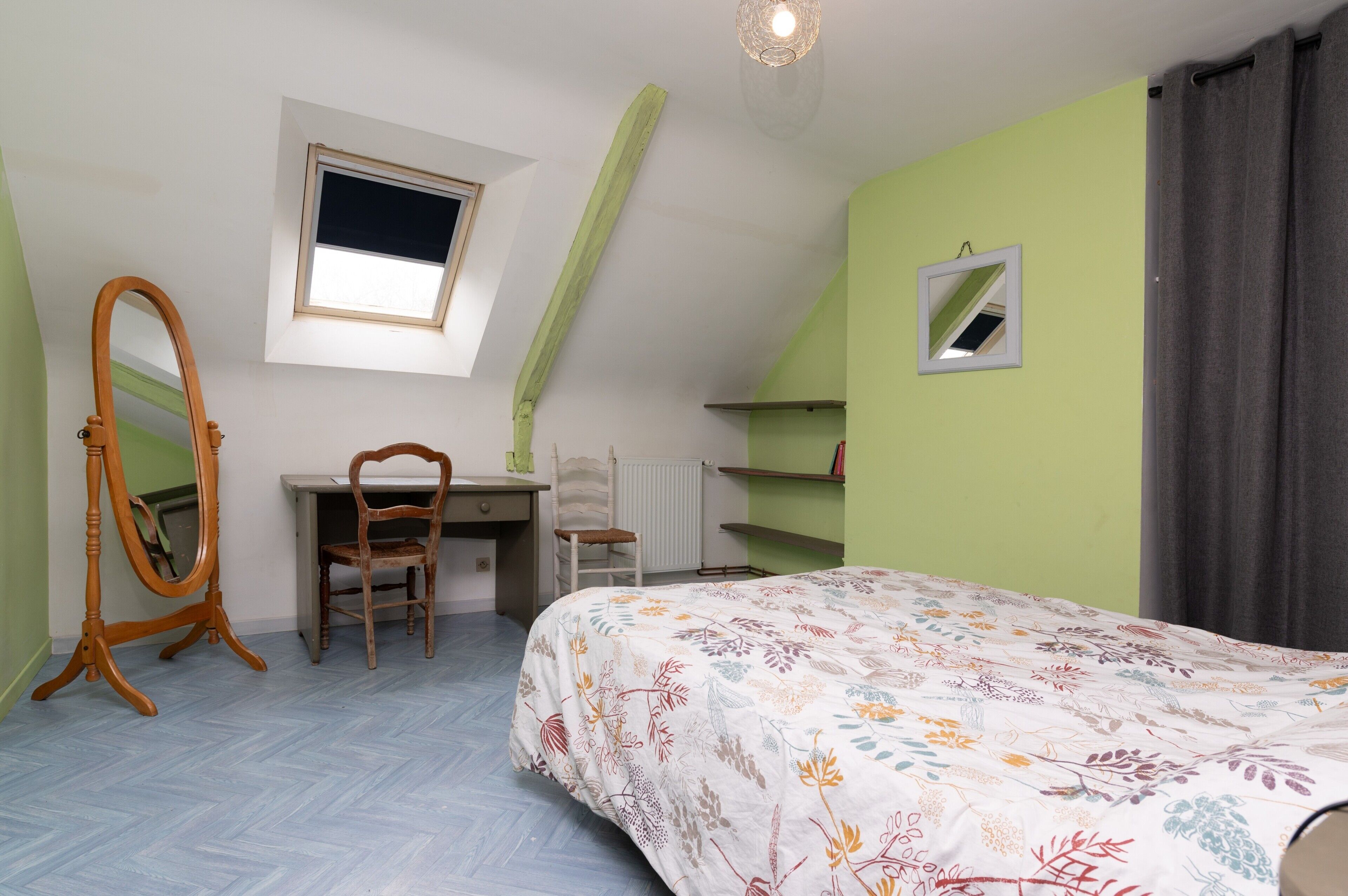 A bedroom with a bed, a desk, a chair, a mirror, a window, and a radiator.