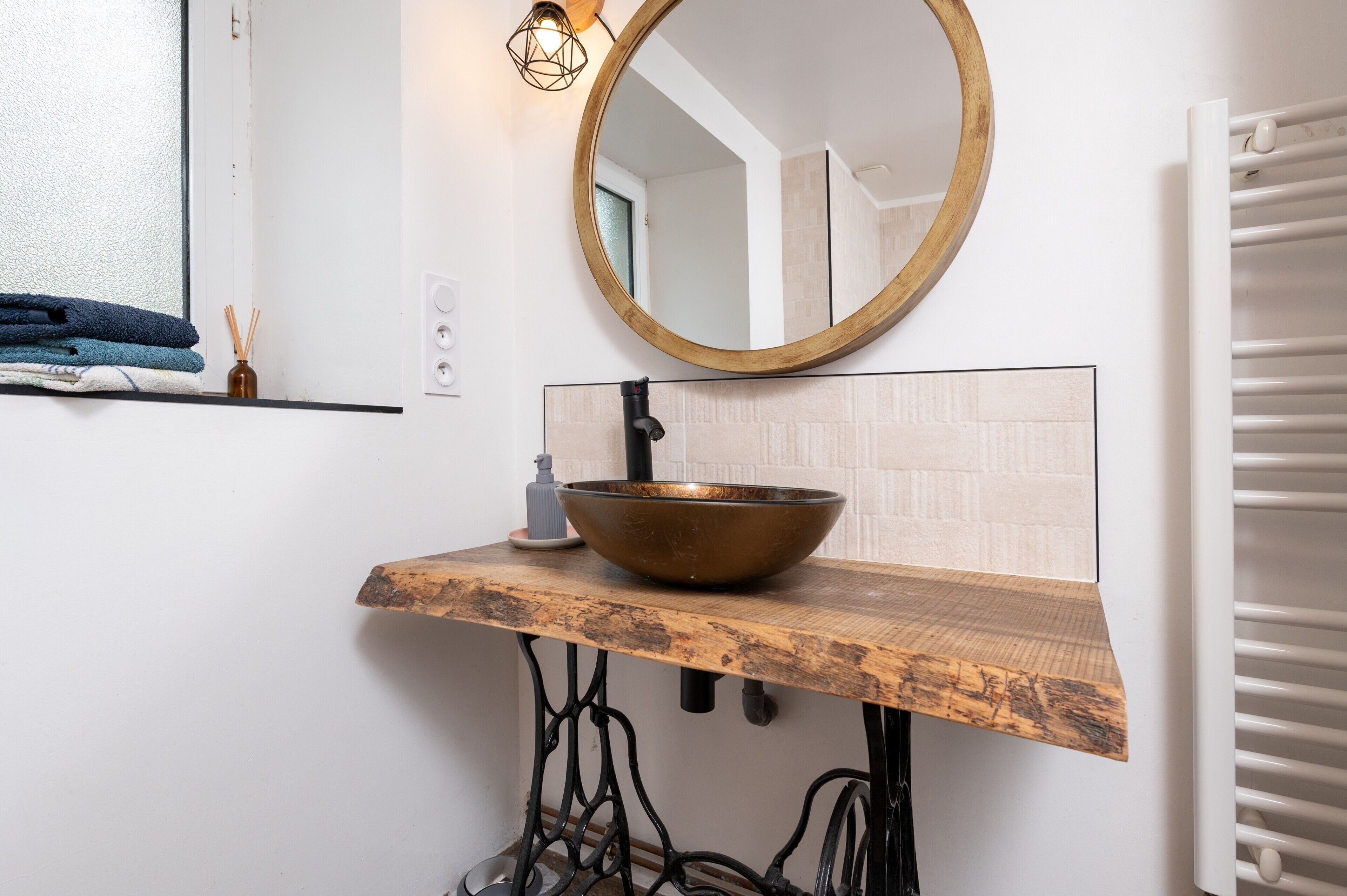 A bathroom with a wooden vanity, a round mirror, a large bowl sink, a towel rack, and a white radiator.
