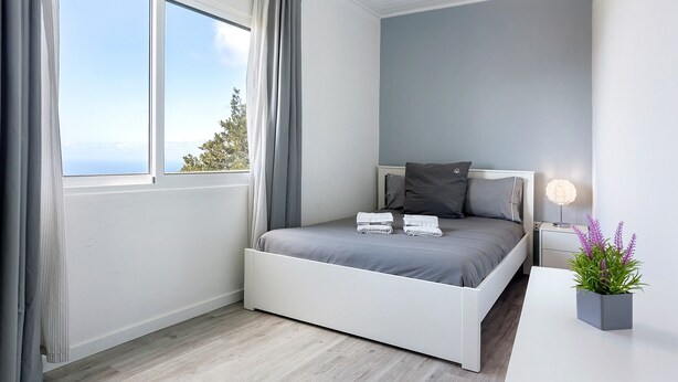 A bedroom with a bed, a bedside table, a lamp, and a window with curtains. - Casa Pereira 2: Perfect for Relaxing and Enjoying Beautiful Sunsets (Ponta do Pargo)