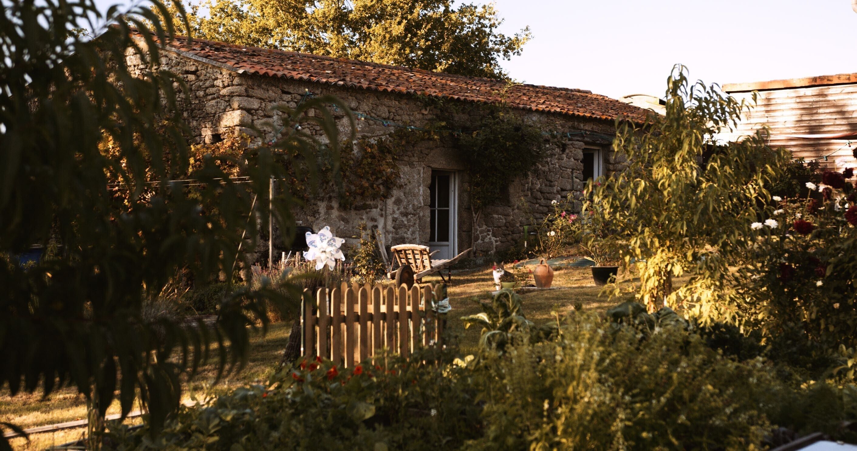 A rustic stone building with a tiled roof, surrounded by lush greenery and a wooden fence.