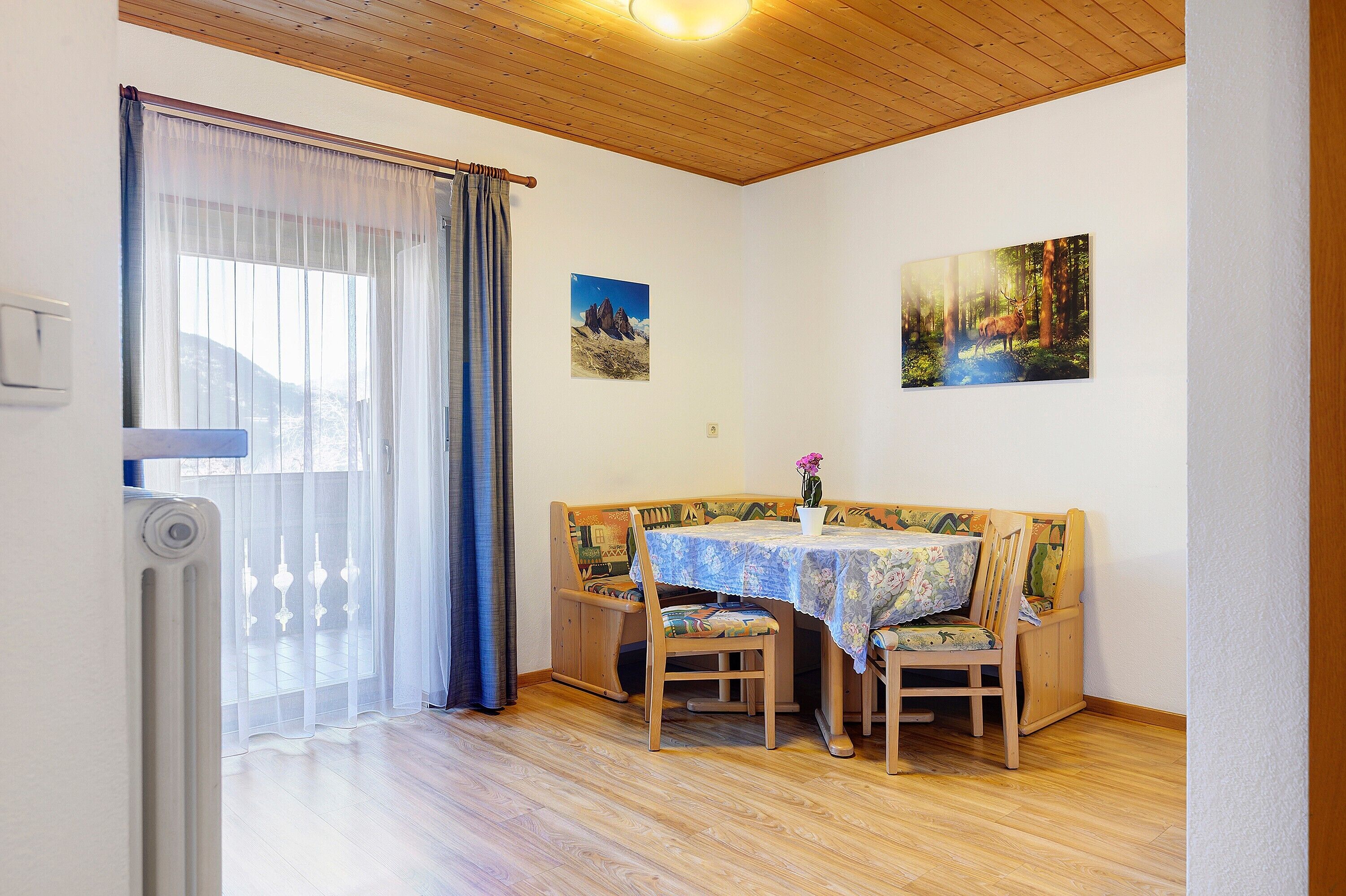 Une pièce avec un plafond en bois, un radiateur, un banc avec des chaises, une table recouverte d’une nappe à motifs floraux et deux tableaux encadrés accrochés au mur.