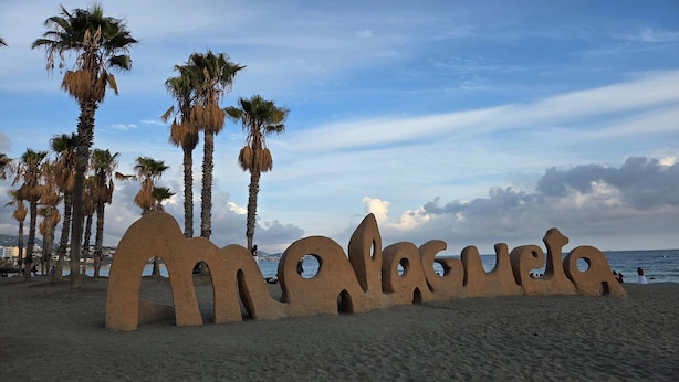 Ein Strand mit einer großen Sandskulptur, die „Amalfi“ buchstabiert, und Palmen im Hintergrund.