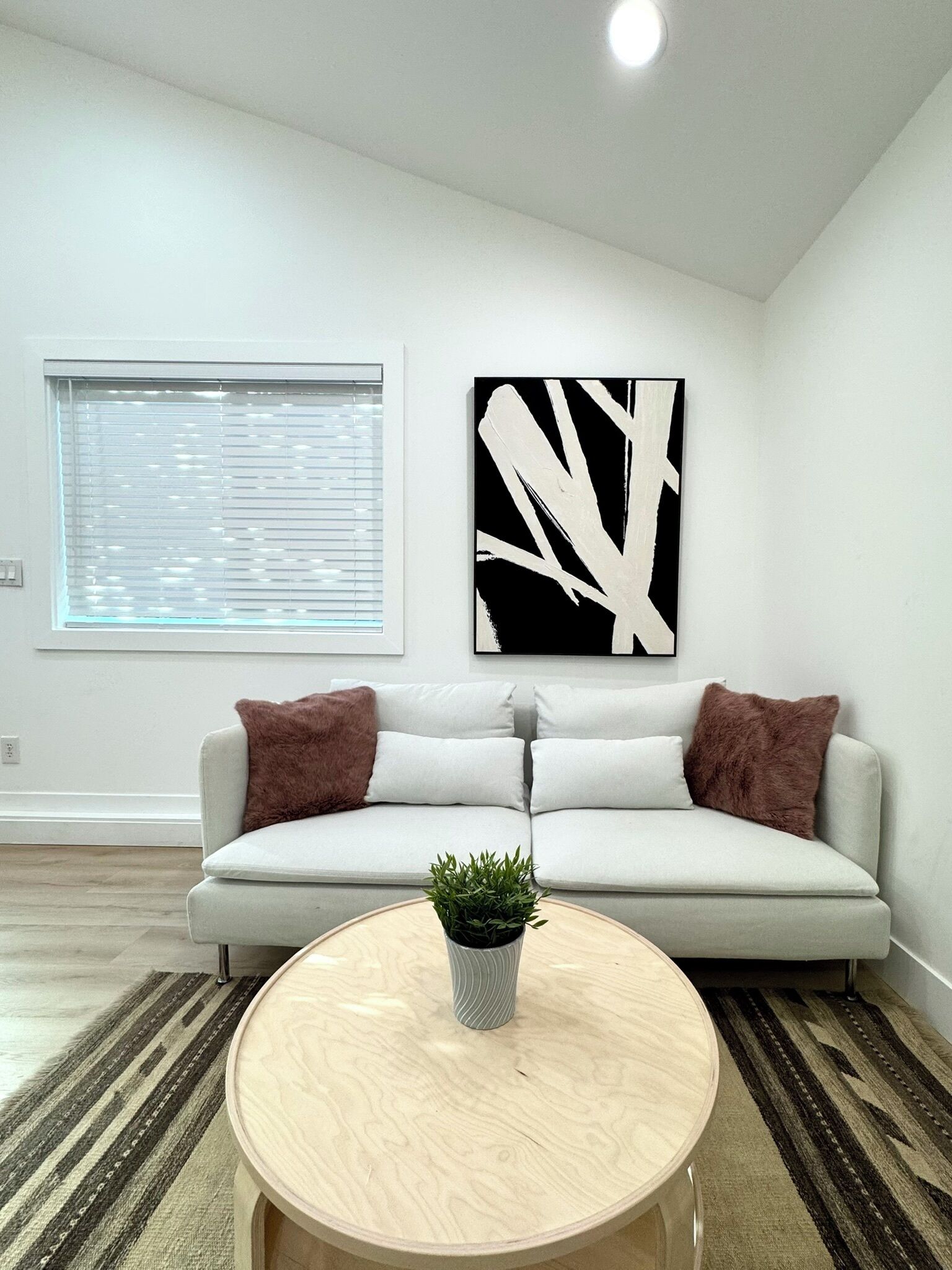A modern living room with a white sofa, two brown cushions, a round wooden coffee table with a plant, and a black and white abstract painting on the wall.