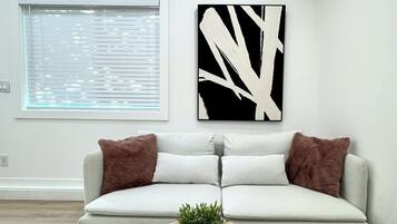 A modern living room with a white sofa, two brown cushions, a round wooden coffee table with a plant, and a black and white abstract painting on the wall.