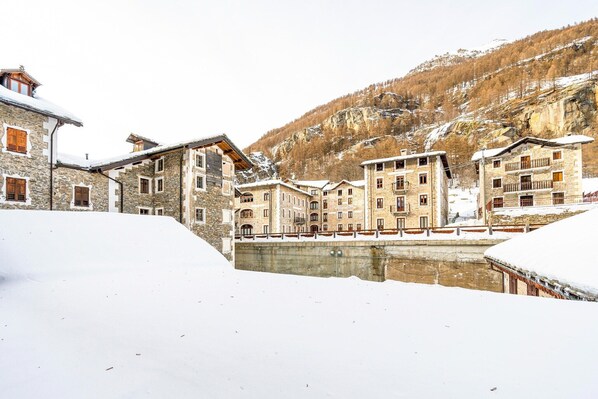 A snow-covered village with stone buildings, a bridge, and a mountain backdrop.