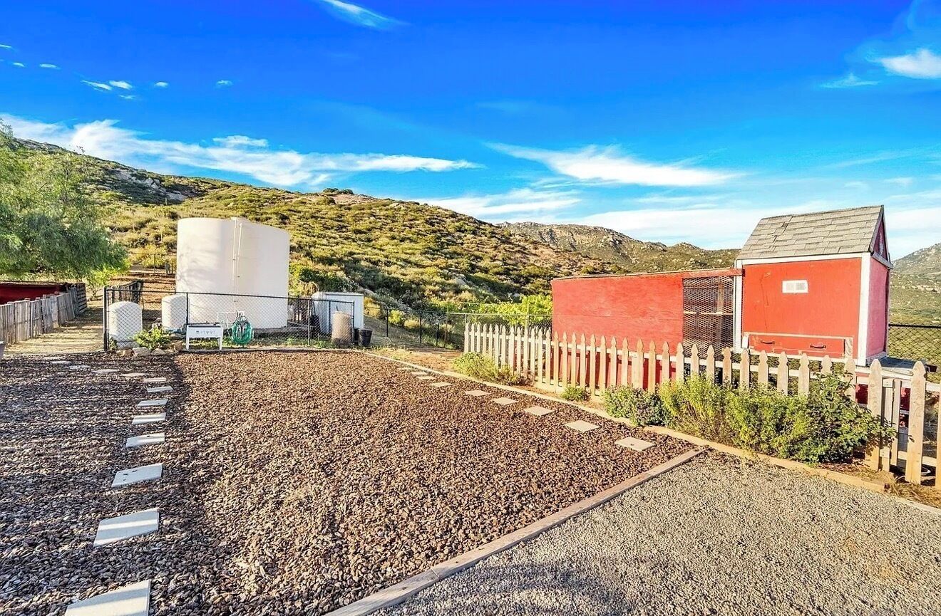 A rural property with a gravel driveway, a red building, and a fenced area with a white fence.