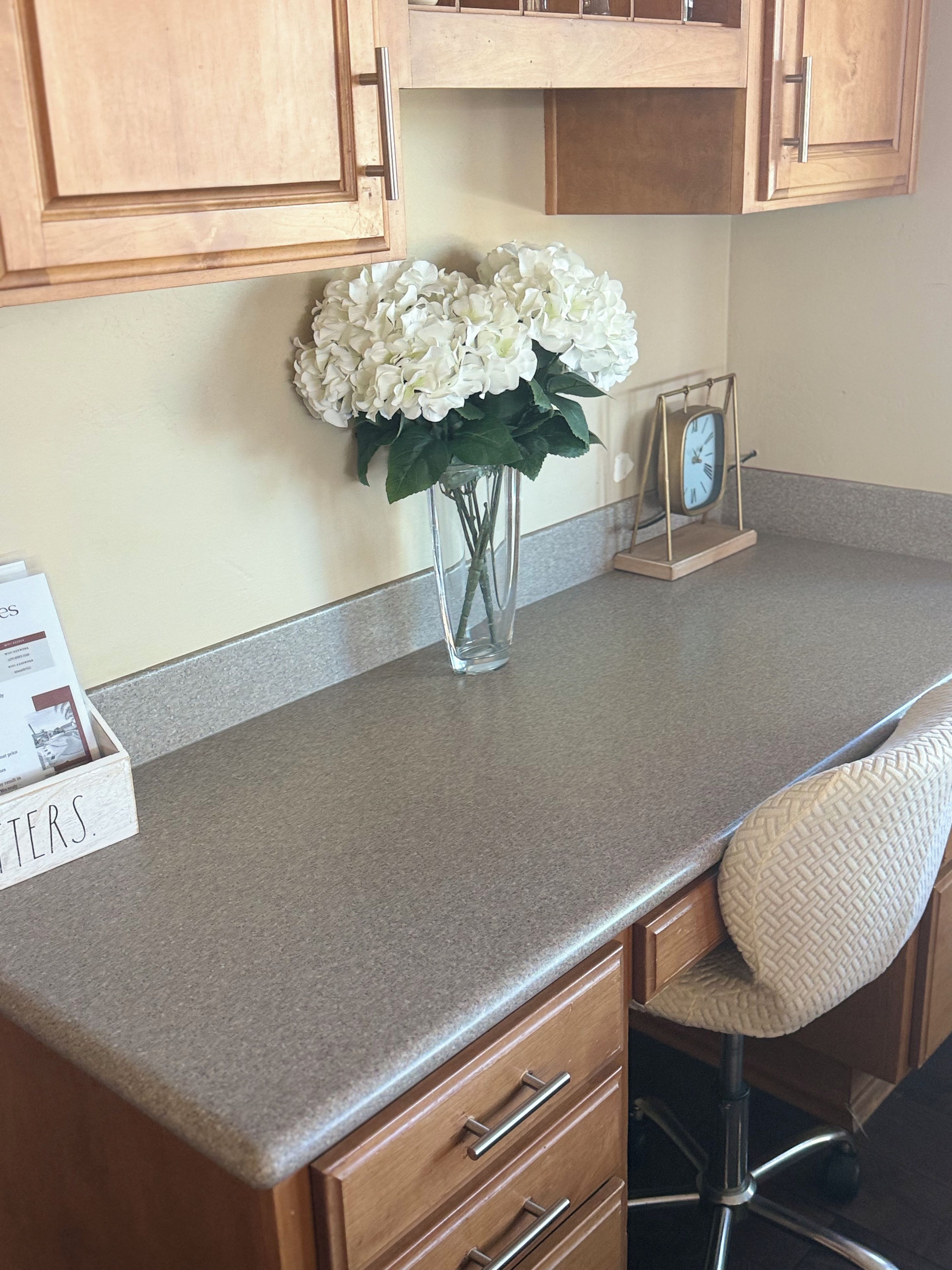 A kitchen counter with a vase of white flowers, a clock, and a wooden box with text.