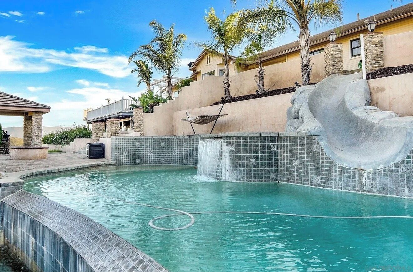 A swimming pool with a waterfall feature, surrounded by palm trees and a stone wall.