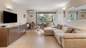 Ein modernes Wohnzimmer mit Flachbildfernseher, einer Couch, einem Esstisch mit Stühlen und einem Balkon mit Blick auf Bäume.