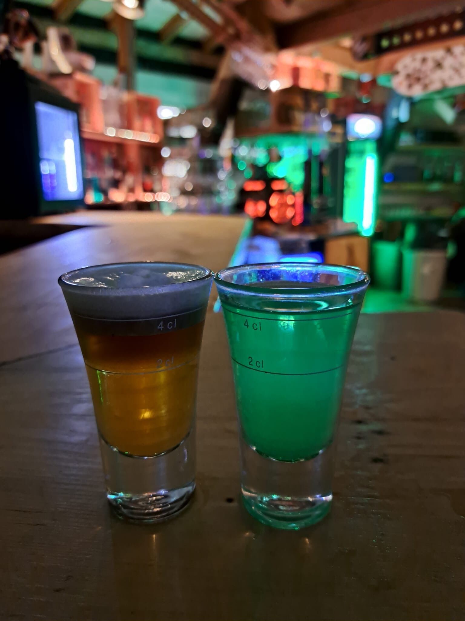 Two shot glasses on a table with measurement markings, in a dimly lit bar setting with colorful lights in the background.