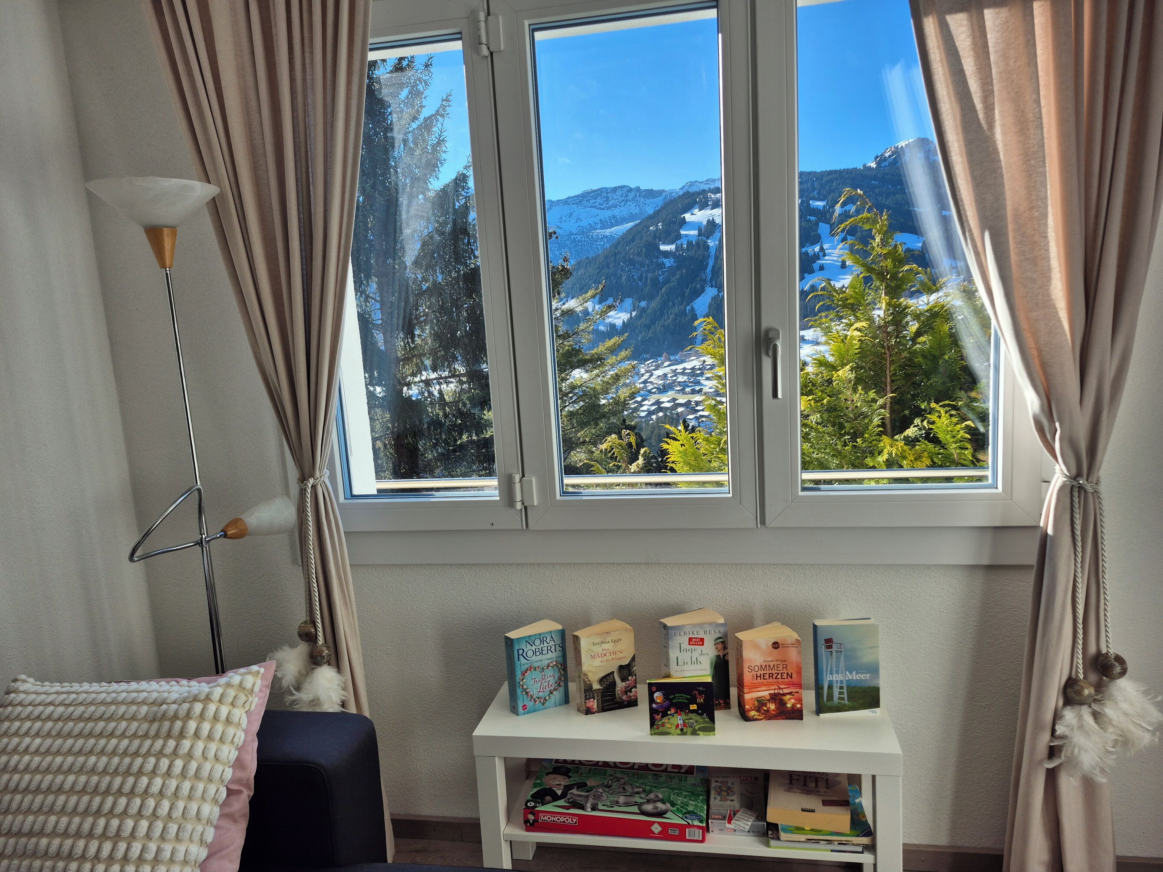 Ein Zimmer mit großem Fenster mit Blick auf eine verschneite Berglandschaft, einem kleinen weißen Regal mit Büchern und einer Lampe.