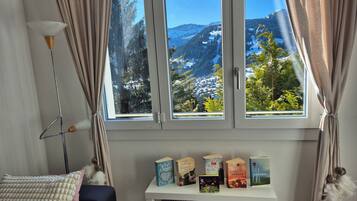 Ein Zimmer mit großem Fenster mit Blick auf eine verschneite Berglandschaft, einem kleinen weißen Regal mit Büchern und einer Lampe.