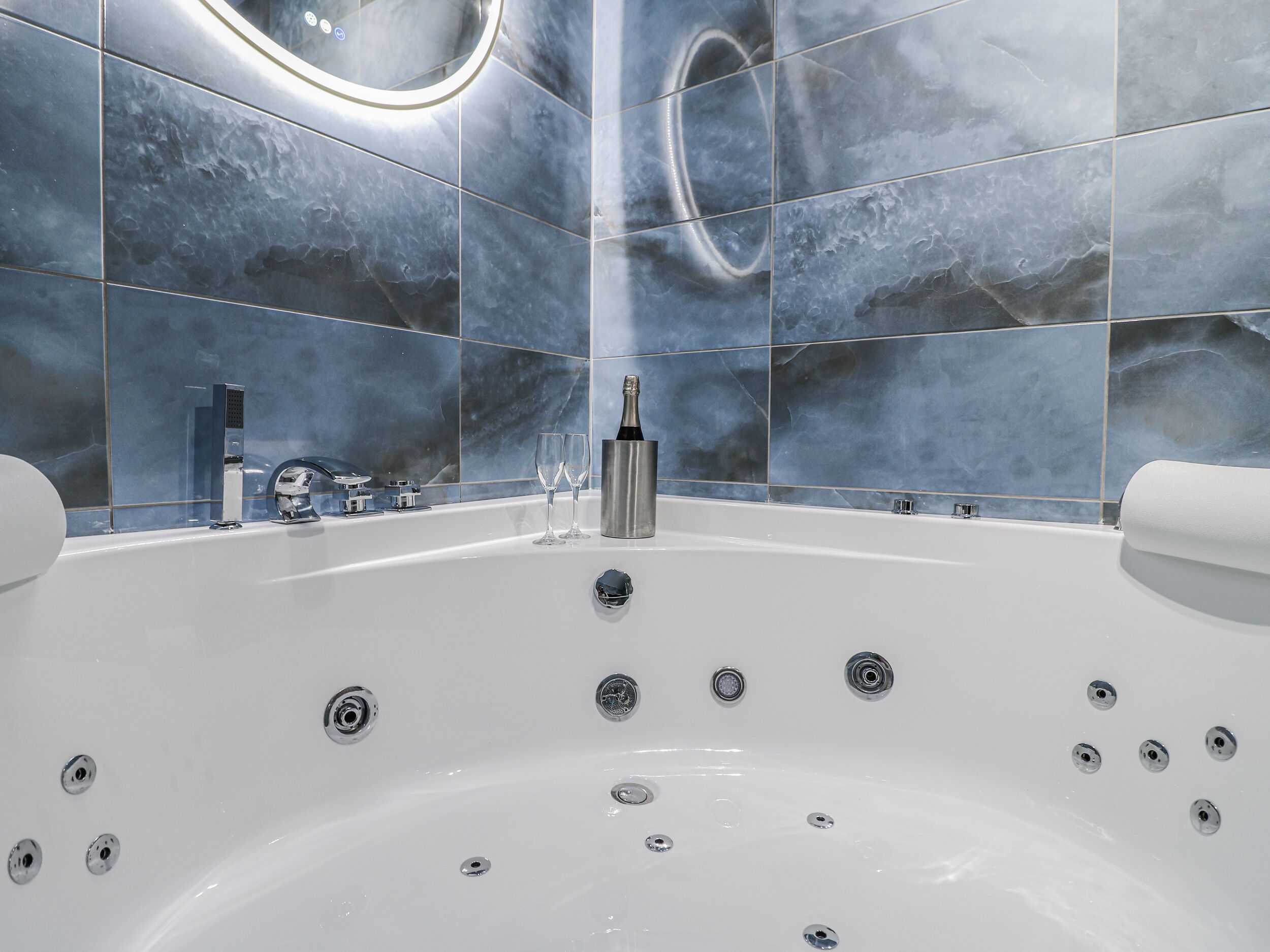 Un baño moderno con una bañera grande, baldosas de mármol y un espejo circular empotrado.