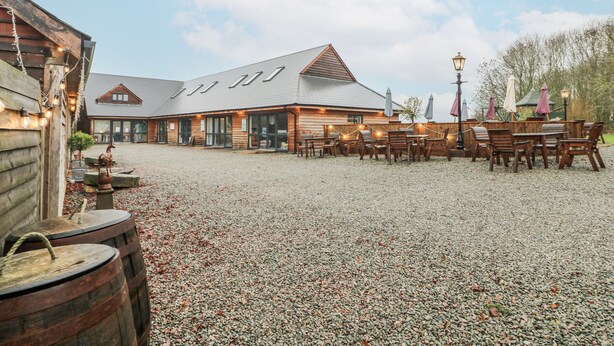 A rustic building with a gravel courtyard, wooden tables and chairs, and a large barrel. - The Walnut Room - Windmill Country Retreats (Melton Mowbray)