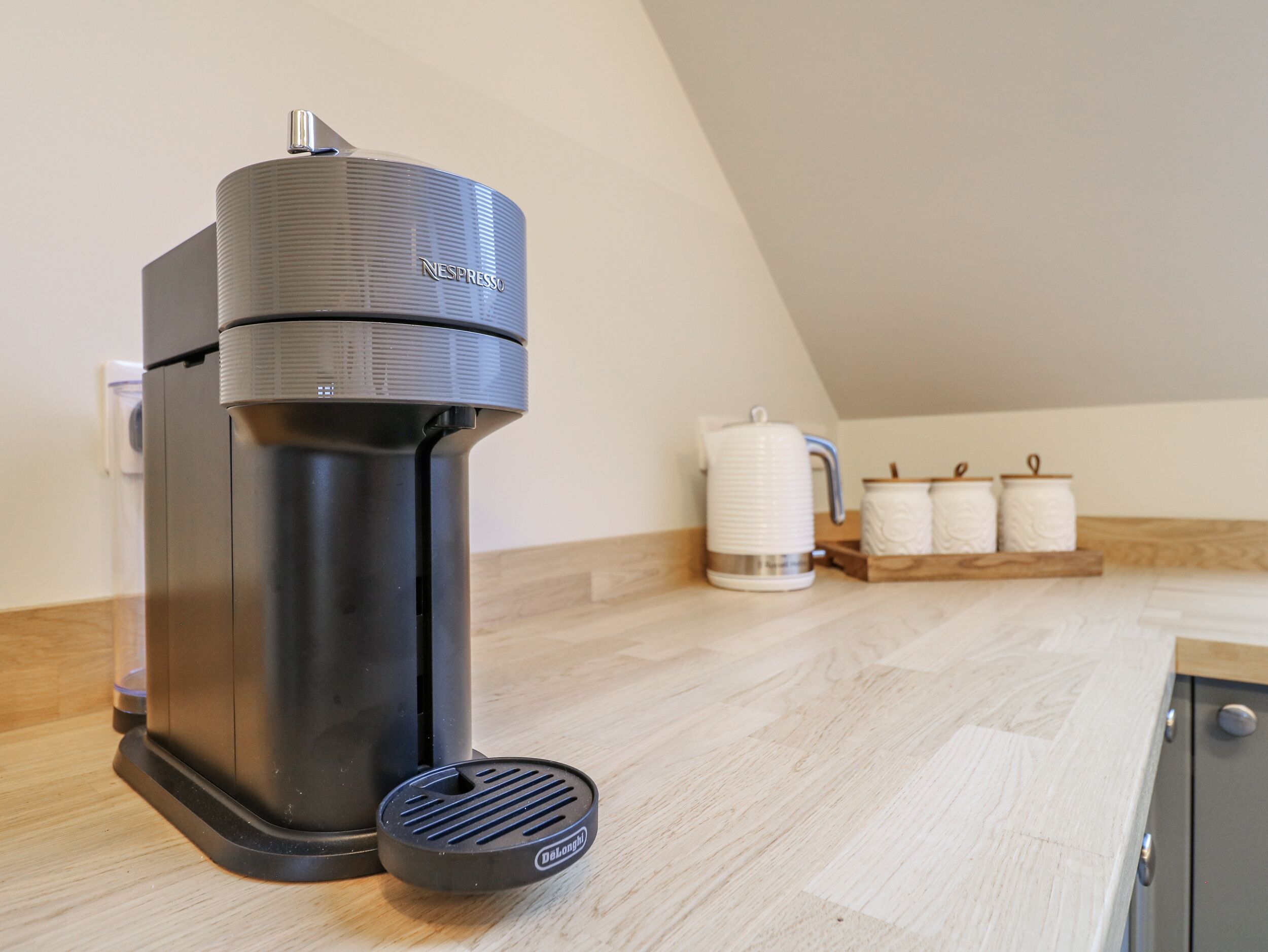 A Nespresso coffee machine on a kitchen counter with a wooden backsplash and a white kettle in the background.