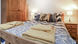 A bedroom with a wooden wardrobe, a bed with patterned bedding, two bedside lamps, and a nightstand with a lamp. - The Walnut Room - Windmill Country Retreats (Melton Mowbray)