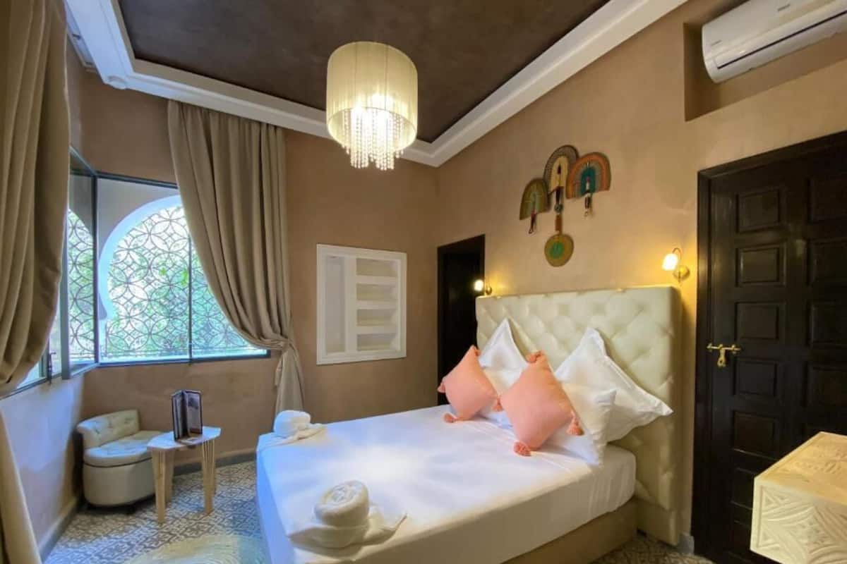 A hotel room with a bed, a small table, a chair, a window with curtains, a wall-mounted air conditioner, and a door.