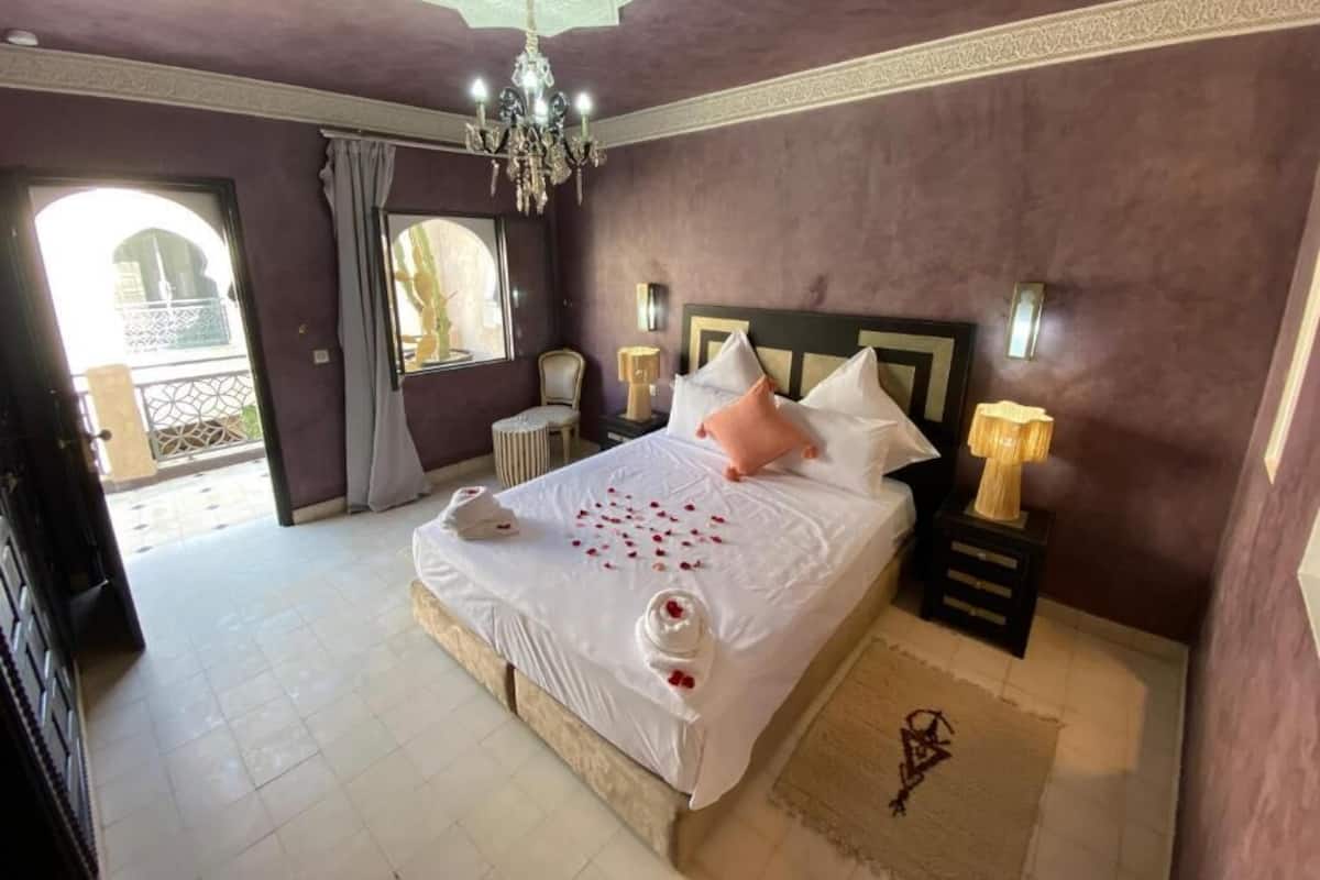 A bedroom with a large bed, a chandelier, a nightstand with a lamp, and a view of a balcony.
