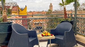 Ein Balkon mit zwei Korbstühlen, einem kleinen Tisch mit Obst und Blick auf Gebäude und einen Turm.
