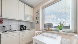 Una cocina con gabinetes blancos, encimera blanca, cafetera y ventana con persianas.