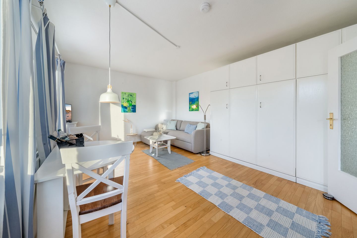 Una sala moderna con sofá, mesa de centro y comedor con silla y mesa. Hay armarios blancos y un ventanal grande.