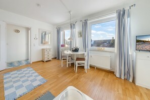 A bright room with wooden flooring, a dining table with chairs, a white cabinet, a radiator, and a TV on the wall. - Cozy 1-room apartment in Hörnum - close to the beach & budget-friendly - (Hörnum (Sylt))