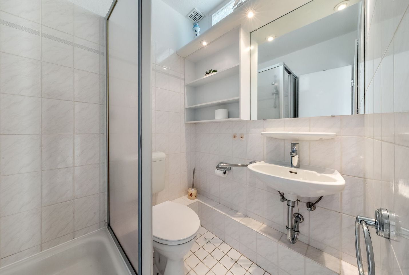 Une salle de bain moderne avec une douche à parois de verre, des toilettes blanches, un lavabo avec un miroir et des murs carrelés.