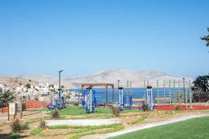 Ein Outdoor-Fitnessbereich mit Trainingsgeräten, einer Holzbank und Blick auf Meer und Berge im Hintergrund.