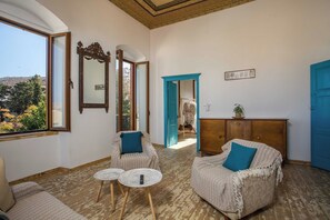 Ein Zimmer mit Holzboden, zwei Sessel mit blauen Kissen, einem kleinen runden Tisch, einer Holzkommode und einem großen Spiegel.