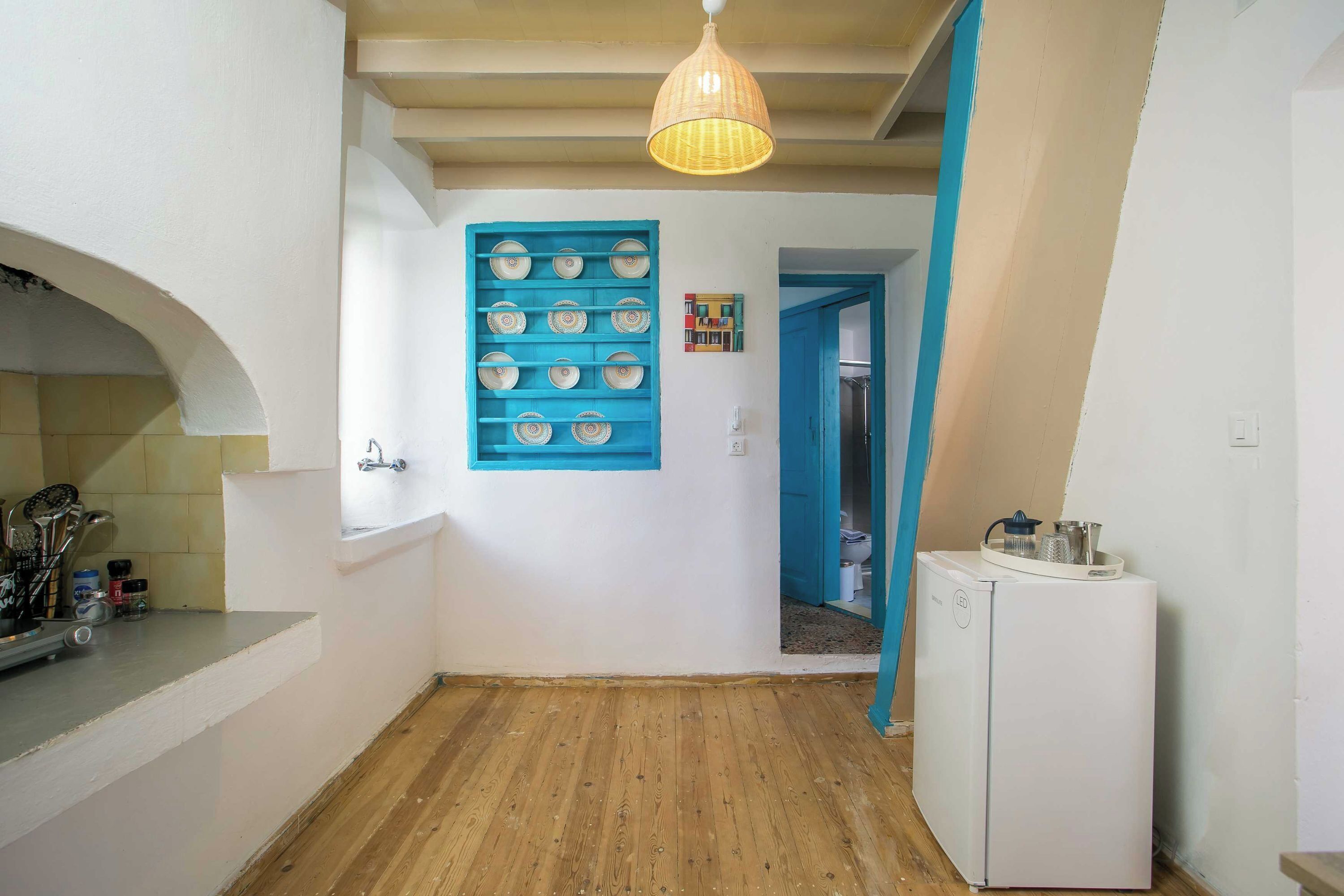 Une cuisine étroite avec un plancher en bois, un réfrigérateur blanc et une étagère bleue fixée au mur, où sont rangés de la vaisselle.