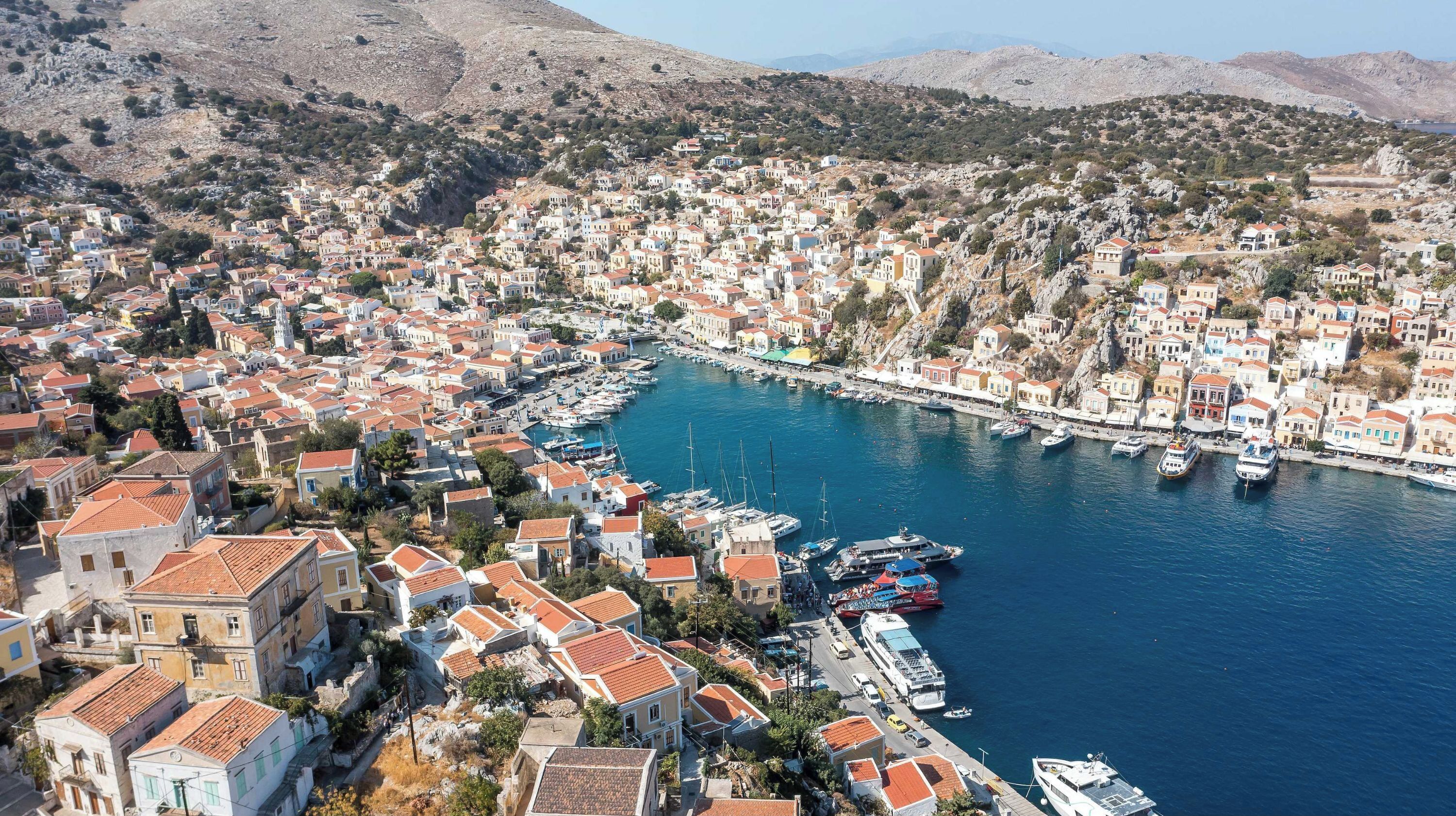 A coastal town with a marina filled with boats, surrounded by densely packed buildings with red-tiled roofs.