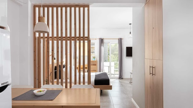 Une pièce moderne et minimaliste, avec une cloison en bois, une table à manger sur laquelle se trouve un saladier de fruits, et un lit agrémenté d’un coussin gris.