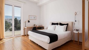 Ein Schlafzimmer mit einem großen Bett, einem Schreibtisch, einem Sessel, einem kleinen Tisch, einer Topfpflanze und Blick auf die Stadt durch das Fenster.