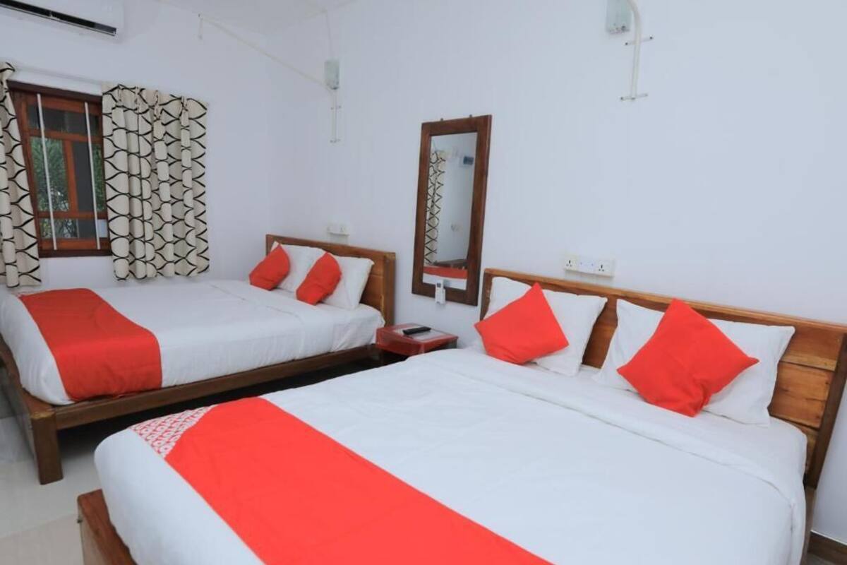 A hotel room with two beds, each with white bedding and red accents, a wooden headboard, a mirror, and a window with patterned curtains.
