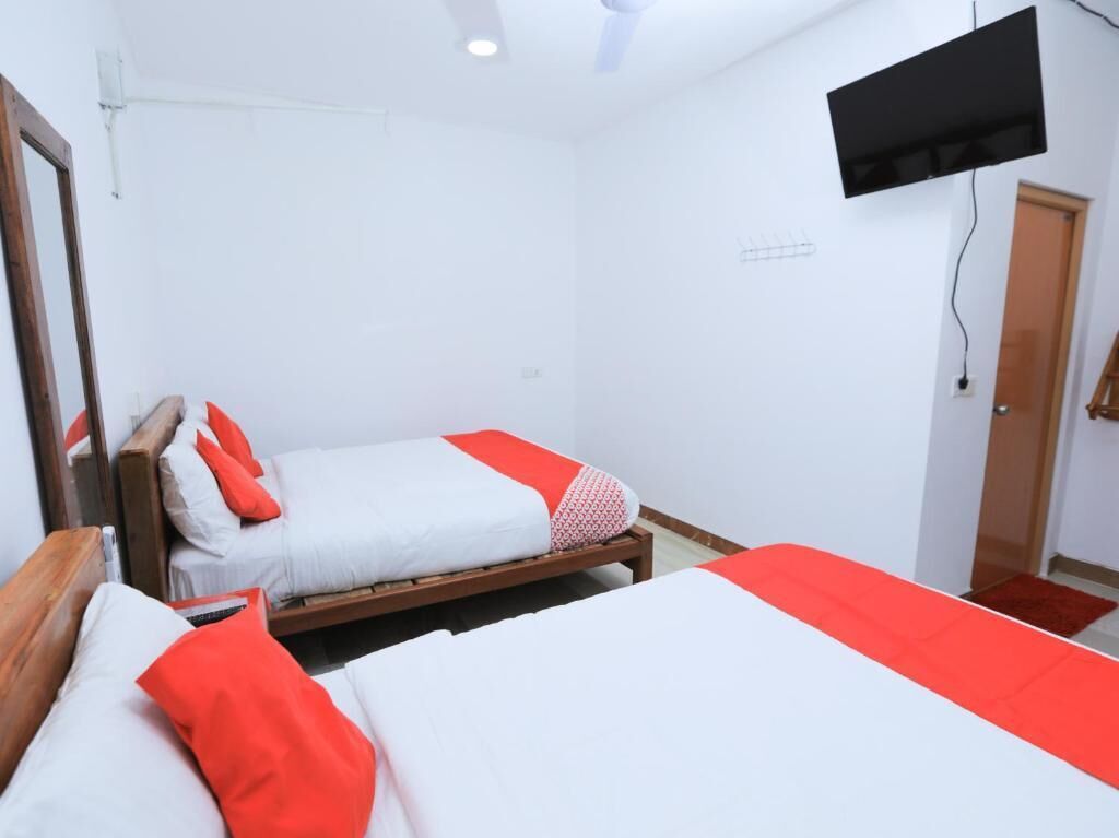 Habitación con dos camas, cada una con ropa blanca y detalles rojos, un televisor montado y una puerta de madera.