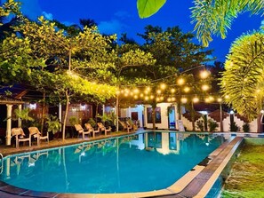 Una piscina con sillas de descanso, luces colgantes y follaje tropical.