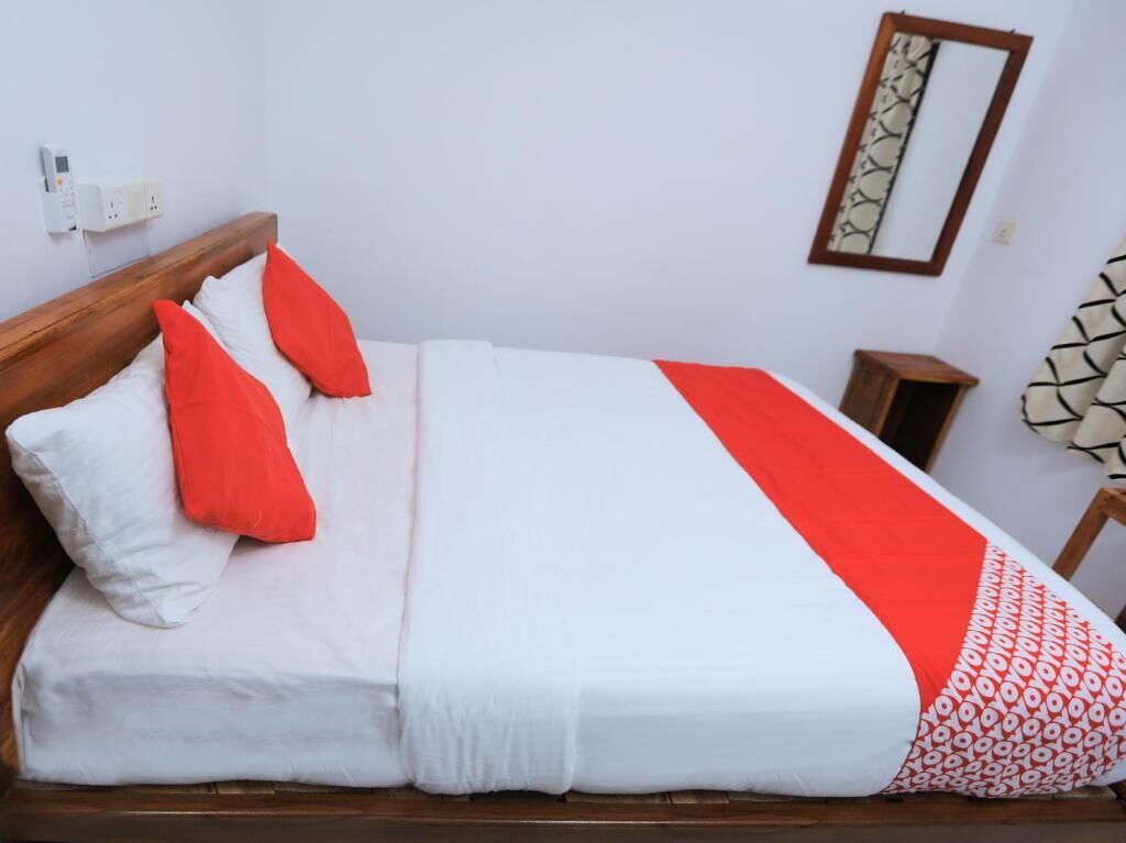 Una cama bien tendida con ropa de cama blanca y roja, un cabecero de madera, una mesita de noche y un espejo montado en la pared.