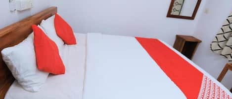 Una cama bien tendida con ropa de cama blanca y roja, un cabecero de madera, una mesita de noche y un espejo montado en la pared.