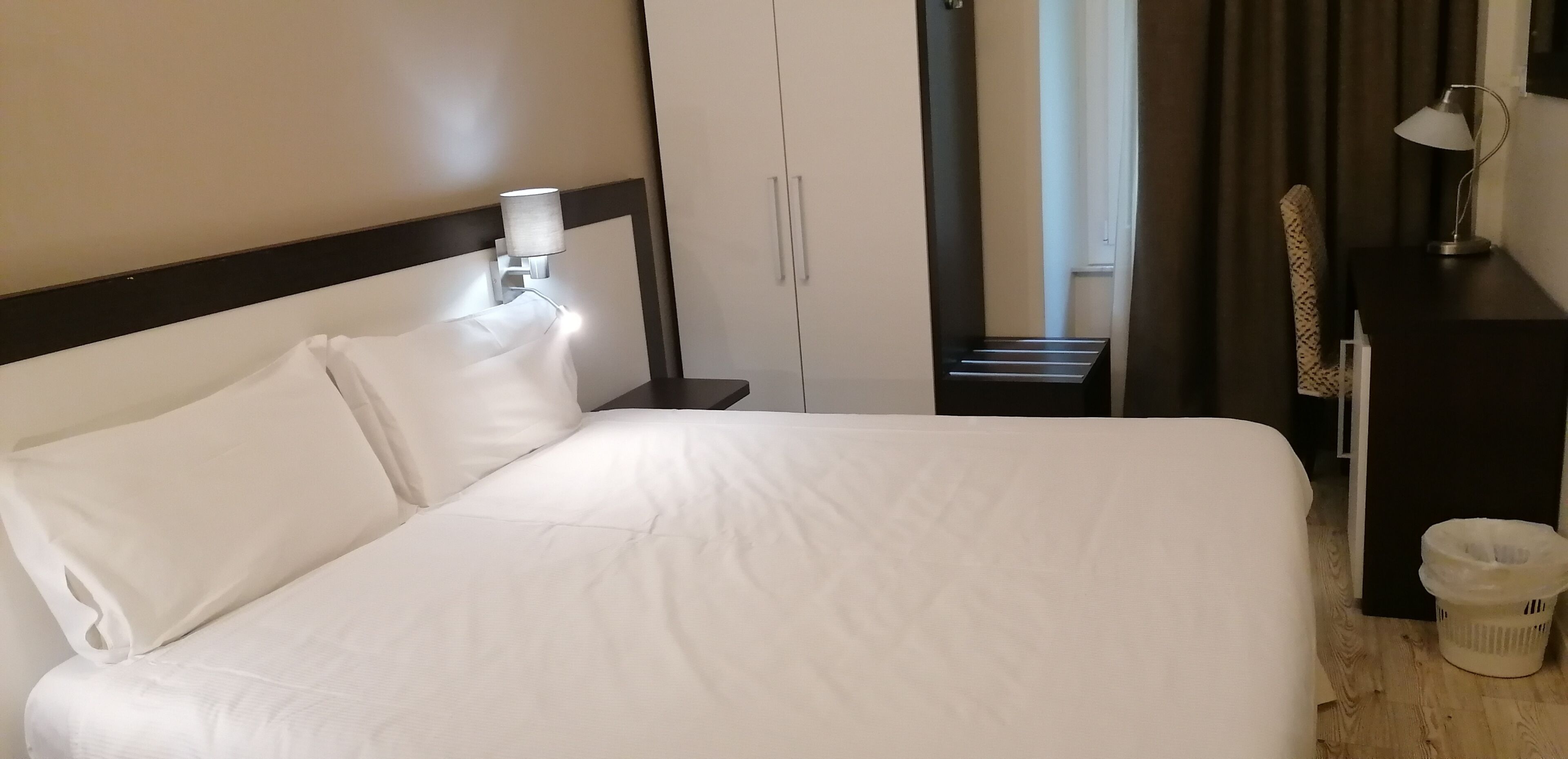 A hotel room with a large bed, a nightstand with a lamp, a wardrobe, and a view of a window with curtains.