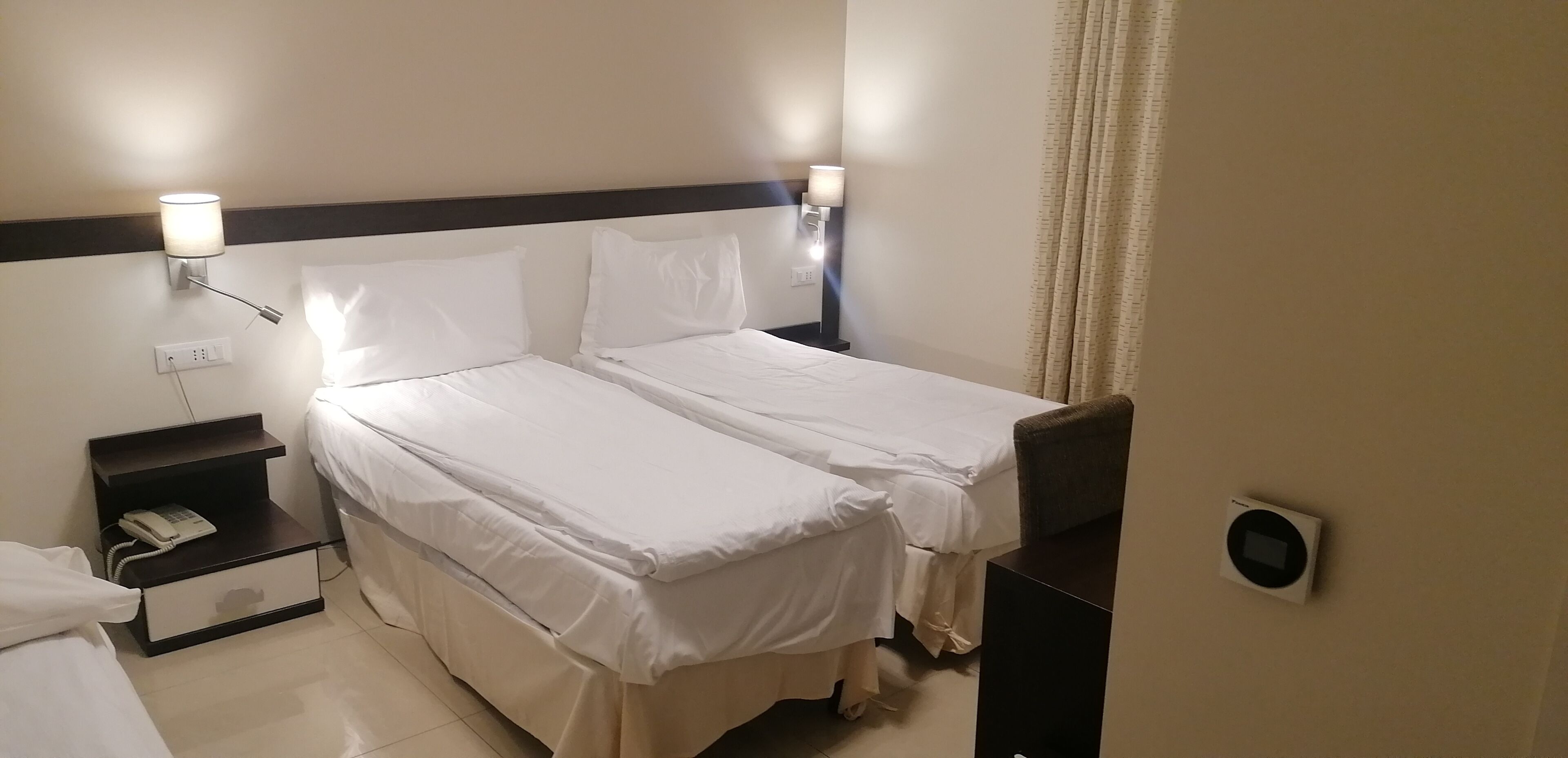 A hotel room with two beds, a bedside table with a phone, and wall-mounted lamps.