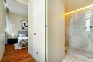 A modern hotel room with a wooden floor, a bed with a checkered bedspread, a nightstand with a lamp, and a marble wall with a wet floor sign. - Riverside Flat Steps from Lisbon’s Attractions (Lisbon)