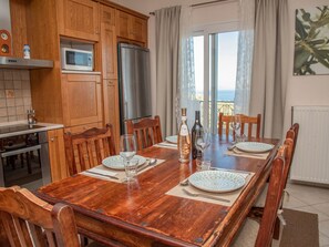 A dining area with a wooden table set for four, a kitchen with wooden cabinets and appliances, and a view through a large window. - Villa Sotiris mit Pool und herrlichem Meerblick by Interhome (Kalami)