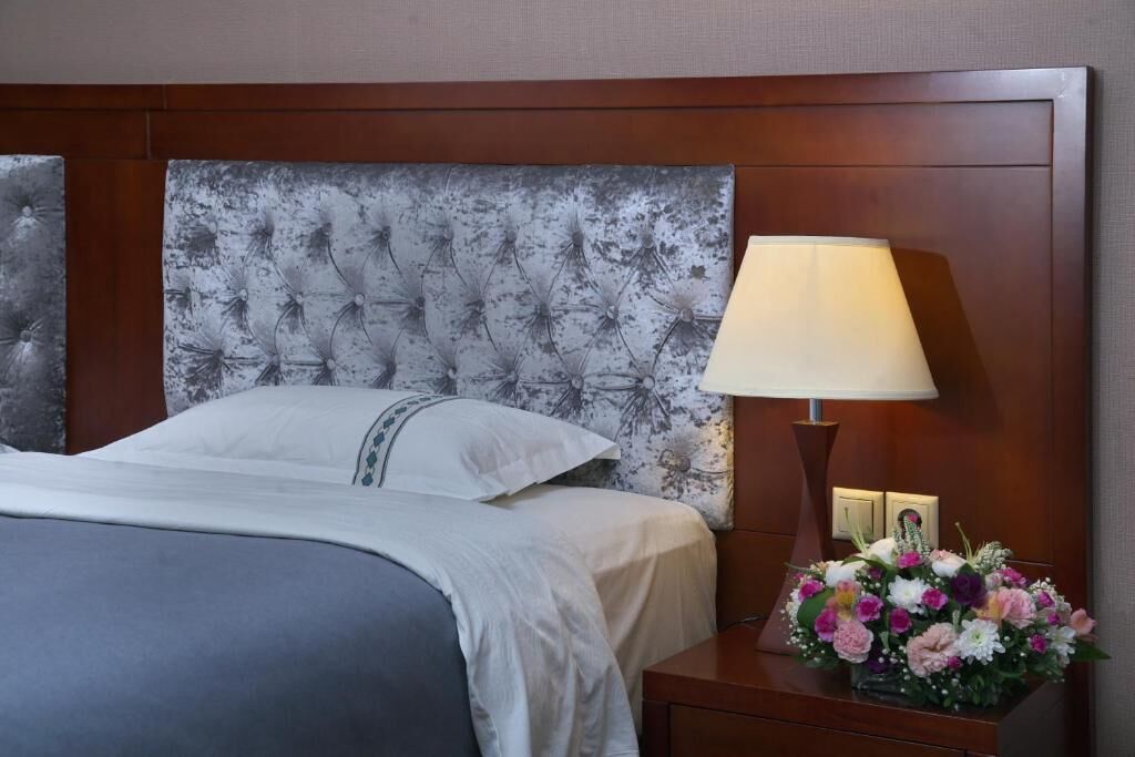 Ein ordentlich bezogenes Bett mit einer grau-weißen Decke, ein Holz-Kopfteil mit Blumenmuster, eine Nachttischlampe und eine Blumenkomposition auf dem Nachttisch.