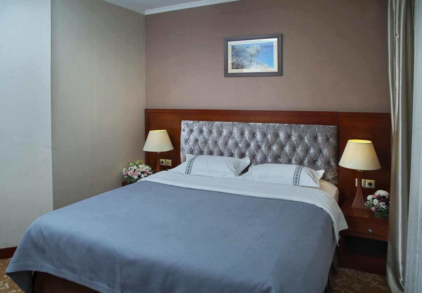 Quarto de hotel com uma cama grande, duas mesas de cabeceira com abajures, um quadro na parede e uma janela com cortinas.