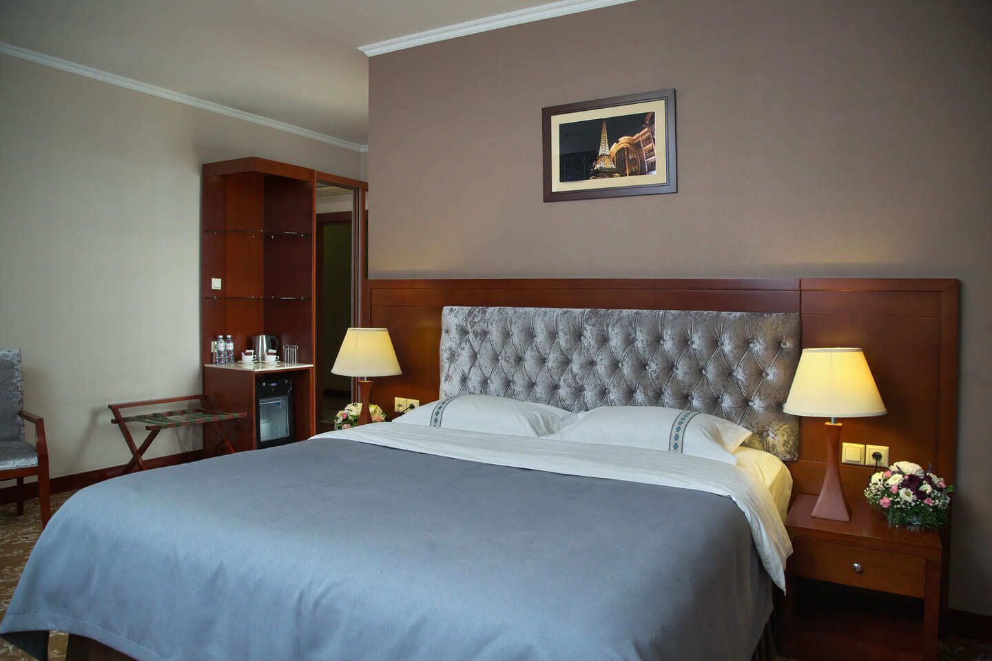 A hotel room with a large bed, two bedside lamps, a nightstand with a vase of flowers, a wooden wardrobe, and a framed picture on the wall.
