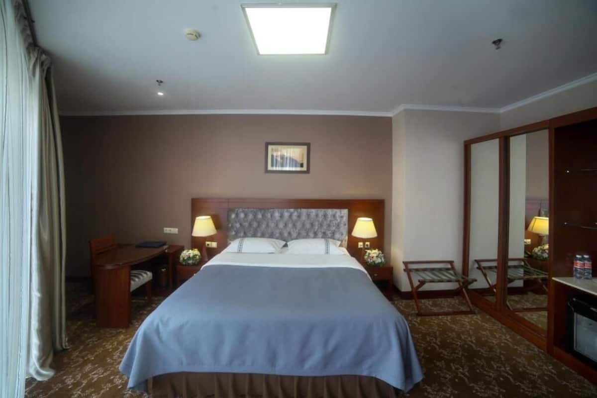 A hotel room with a large bed, two bedside tables with lamps, a small desk, a chair, a mirror, and a window with curtains.