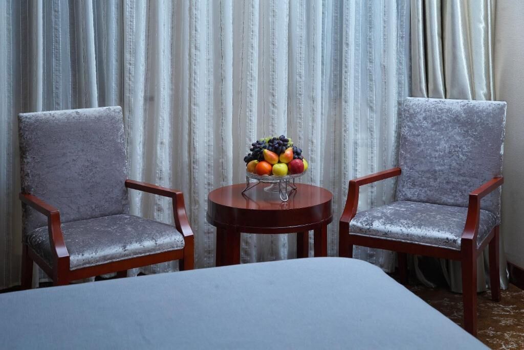 Una habitación con dos sillones tapizados grises, una mesita redonda de madera con fruteros, y una cama con cubierta gris.