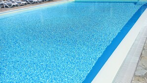 Una piscina trasparente con bordo bianco e area solarium piastrellata con lettini.