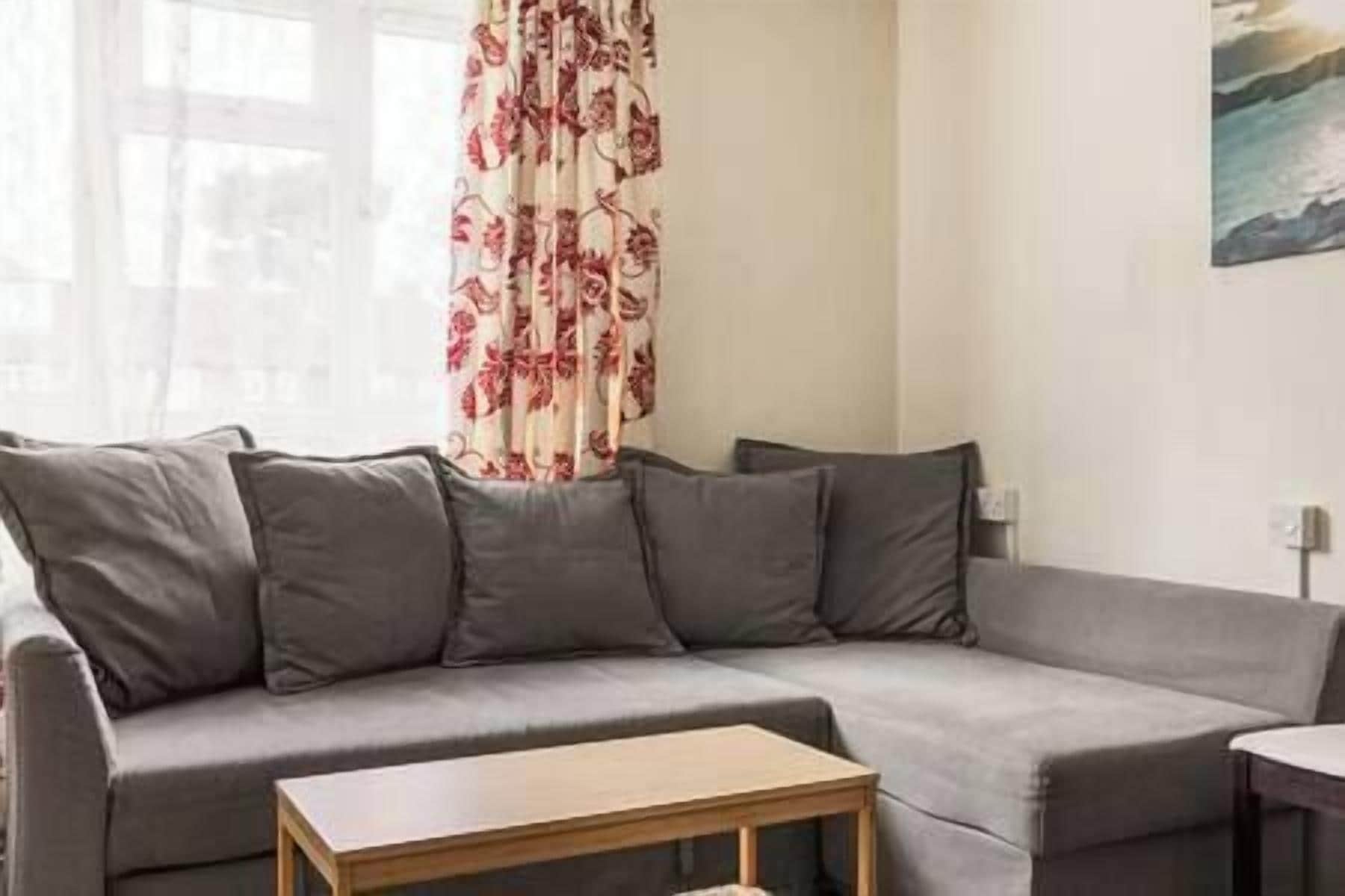 Ein graues Sofa mit mehreren Kissen, ein kleiner Holztisch und ein Fenster mit blumigen Vorhängen.