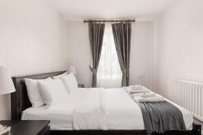 A neatly made bed with white linens and a gray throw blanket, a bedside lamp, and a window with gray curtains. - New! Inviting 1BR Flat by Richmond Park w/Parking (Greater London)